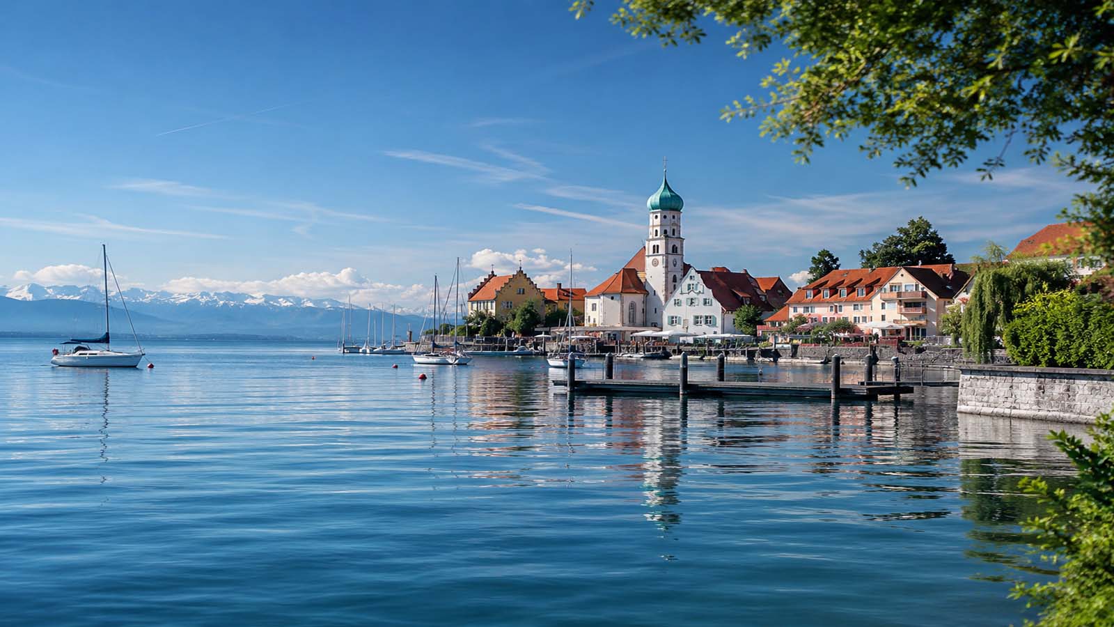 Blick auf die Halbinsel Wasserburg am Bodensee mit Kirche, Hafen, Segelbooten und Alpen im Hintergrund.